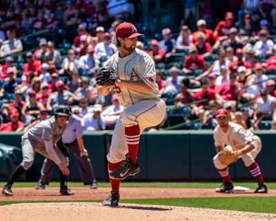 A baseball pitcher is in the middle of his windup on a sunny day during a game. He is wearing a uniform with red accents and a cap, and there is a crowd in the stands, most likely fans, watching intently. An opposing player is positioned nearby, possibly getting ready to run.