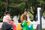 A young girl and a young man are sitting on colorful playground rides shaped like animals. The girl is wearing a pink sweater and skirt with black and white striped socks, and she is looking away while covering her face with a hand. The man, wearing a brown jacket and carrying a backpack, is looking at the girl. The background is a park with green trees.