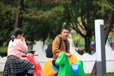 A young girl and a young man are sitting on colorful playground rides shaped like animals. The girl is wearing a pink sweater and skirt with black and white striped socks, and she is looking away while covering her face with a hand. The man, wearing a brown jacket and carrying a backpack, is looking at the girl. The background is a park with green trees.