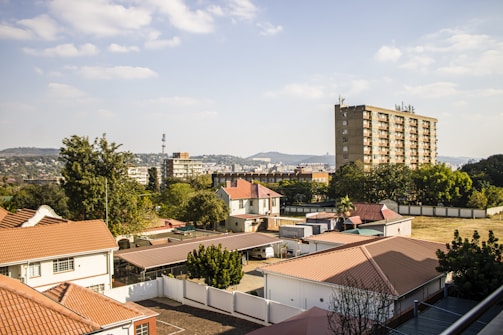 A residential area with multiple houses featuring red and brown roofs is visible. In the background, a tall apartment building is situated surrounded by trees and other smaller buildings. The skyline includes distant hills under a partly cloudy sky.
