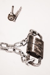 A padlock securely fastened with a chain, placed next to a set of keys on a white background. The metal surface of the padlock and chain reflects light, giving a shiny appearance.