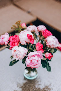 A close-up of an elegant floral arrangement featuring deep burgundy roses, soft blush peonies, and delicate greenery in a sleek glass vase.