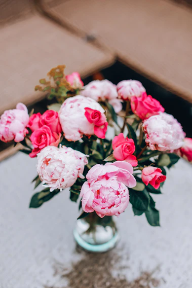 A close-up of an elegant floral arrangement featuring deep burgundy roses, soft blush peonies, and delicate greenery in a sleek glass vase.