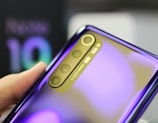A close-up view of a person's hand holding a smartphone showing the rear camera setup. The phone features a reflective, shiny surface with a gradient finish that shifts from purple to a lighter hue. The camera module has four lenses arranged vertically, with text indicating 'AI QUAD CAMERA' next to them.