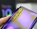 A close-up view of a person's hand holding a smartphone showing the rear camera setup. The phone features a reflective, shiny surface with a gradient finish that shifts from purple to a lighter hue. The camera module has four lenses arranged vertically, with text indicating 'AI QUAD CAMERA' next to them.