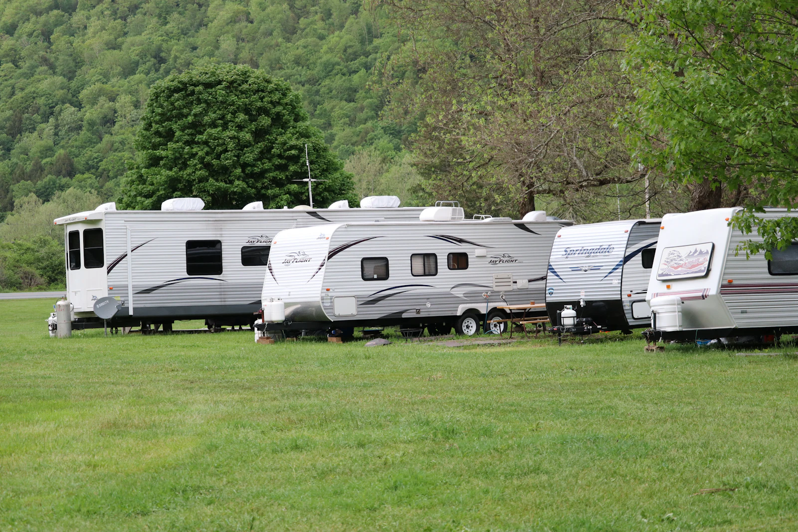 A row of travel trailers parked on grass campsites with utility posts and propane tanks beside them and a wooded hillside behind