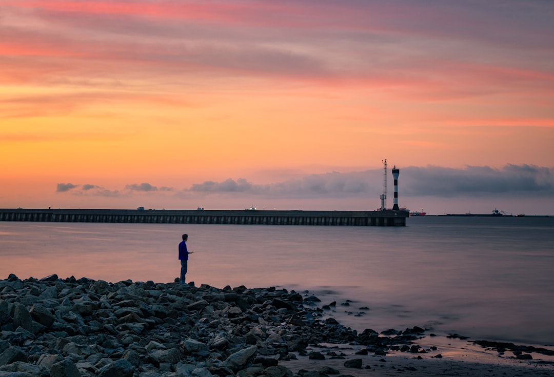 person standing on rocky shore near bridge during sunset