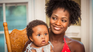 A smiling mother holding a nursing pillow in a sunlit living room.