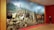 A large, detailed painting depicts a bustling pastoral scene with numerous animals including cows and sheep, and people dressed in rural attire. In the foreground, horseback riders are prominent, accompanied by dogs. The backdrop features a vivid landscape with a town and mountainous terrain.