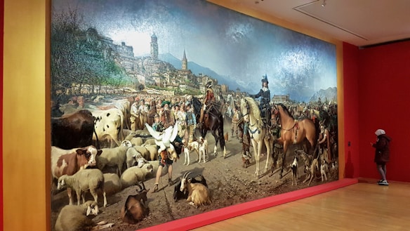 A large, detailed painting depicts a bustling pastoral scene with numerous animals including cows and sheep, and people dressed in rural attire. In the foreground, horseback riders are prominent, accompanied by dogs. The backdrop features a vivid landscape with a town and mountainous terrain.