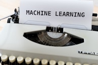 A vintage typewriter with a sheet of paper on which the words 'MACHINE LEARNING' are typed in bold. The typewriter appears to be an older model with black keys and a white body, placed on a wooden surface.