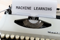 A vintage typewriter with a sheet of paper on which the words 'MACHINE LEARNING' are typed in bold. The typewriter appears to be an older model with black keys and a white body, placed on a wooden surface.