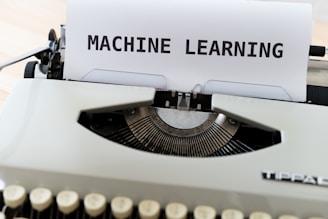 A vintage typewriter with a sheet of paper on which the words 'MACHINE LEARNING' are typed in bold. The typewriter appears to be an older model with black keys and a white body, placed on a wooden surface.