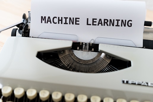 A vintage typewriter with a sheet of paper on which the words 'MACHINE LEARNING' are typed in bold. The typewriter appears to be an older model with black keys and a white body, placed on a wooden surface.