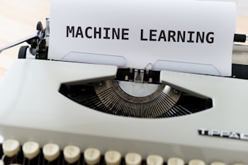 A vintage typewriter with a sheet of paper on which the words 'MACHINE LEARNING' are typed in bold. The typewriter appears to be an older model with black keys and a white body, placed on a wooden surface.