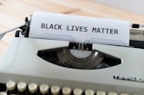 A vintage typewriter with a piece of paper being typed on, reading 'BLACK LIVES MATTER' in uppercase letters. The typewriter is situated on a light wooden surface, with focus on the text and the typewriter keys partially visible.