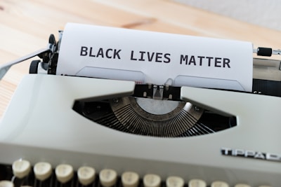 A vintage typewriter with a piece of paper being typed on, reading 'BLACK LIVES MATTER' in uppercase letters. The typewriter is situated on a light wooden surface, with focus on the text and the typewriter keys partially visible.