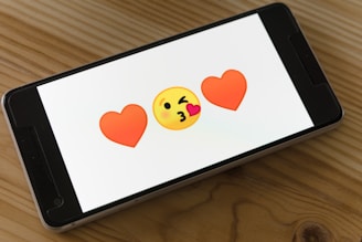 A smartphone displays two red heart emojis flanking a yellow face blowing a kiss on a wooden table.
