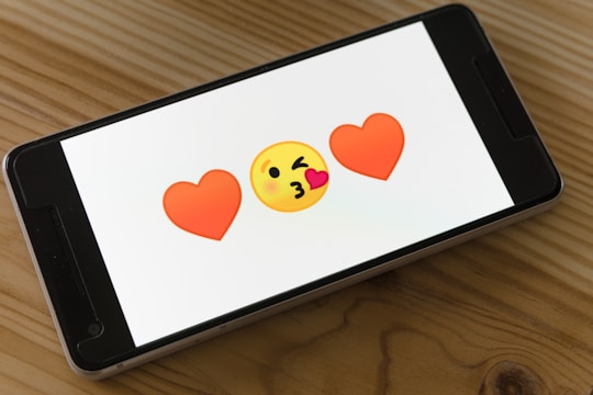 A smartphone displays two red heart emojis flanking a yellow face blowing a kiss on a wooden table.