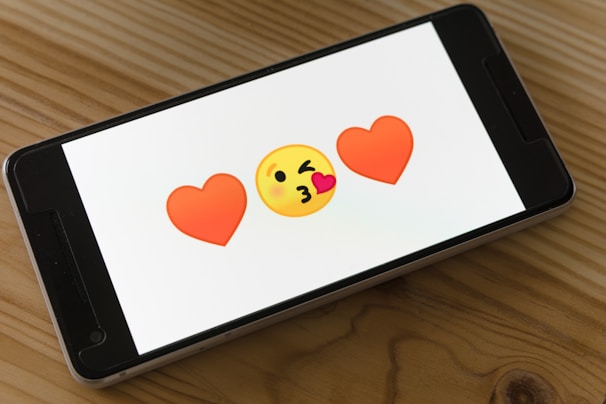 A smartphone displays two red heart emojis flanking a yellow face blowing a kiss on a wooden table.
