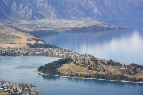 A panoramic view of a lakeside town surrounded by mountains. The foreground features a body of water with a shoreline bordered by trees. The town consists of a cluster of residential buildings nestled among patches of greenery. Beyond the town, tall mountains rise with rocky surfaces and patches of vegetation.