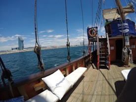 A view from the deck of a wooden sailing ship, showing the expansive blue sea and a clear sky. A large modern building can be seen in the distance on the shoreline. Life rafts and ropes are present on the ship. Cushioned seating with branded pillows is visible, suggesting a leisure cruise or tour.