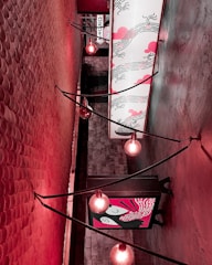 The image features a narrow, stylish indoor alleyway with walls covered in textured patterns. Warm red lighting from exposed bulb string lights creates an inviting and artistic ambiance. There are two rectangular art panels with decorative designs on the wall, featuring abstract patterns and Asian-inspired motifs. The ceiling is adorned with a custom backlit artwork panel designed with flowing and wave-like patterns.