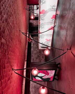 The image features a narrow, stylish indoor alleyway with walls covered in textured patterns. Warm red lighting from exposed bulb string lights creates an inviting and artistic ambiance. There are two rectangular art panels with decorative designs on the wall, featuring abstract patterns and Asian-inspired motifs. The ceiling is adorned with a custom backlit artwork panel designed with flowing and wave-like patterns.