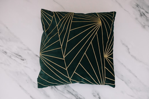A dark green pillow featuring an intricate pattern of golden lines creating geometric shapes. The pillow is placed on a light grey marble surface with subtle veins running through it.