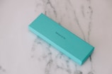 A rectangular turquoise box is lying on a white marble surface with subtle gray veining. The box is labeled with 'TIFFANY & CO.' in black text.