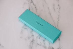A rectangular turquoise box is lying on a white marble surface with subtle gray veining. The box is labeled with 'TIFFANY & CO.' in black text.