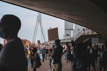 A large group of people gathered under a bridge, some holding signs with messages advocating for social justice and equality. The crowd includes individuals of diverse backgrounds, many wearing masks. The surrounding urban environment is visible, including a modern bridge structure and tall buildings.