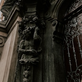 An intricate stone carving of a muscular figure holding an object, possibly a horn, is set into the facade of a building. The surrounding architecture features ornate scrollwork and detailed ironwork on a nearby door, creating an impression of elegance and age.