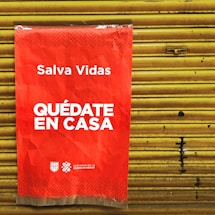 A bright red poster with white text that says 'Salva Vidas Quédate En Casa' is attached to a yellow metal shutter. The message emphasizes staying at home to save lives. The bottom has logos representing the government of Mexico City.
