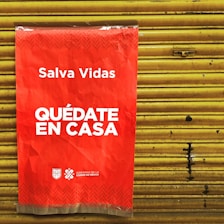 A bright red poster with white text that says 'Salva Vidas Quédate En Casa' is attached to a yellow metal shutter. The message emphasizes staying at home to save lives. The bottom has logos representing the government of Mexico City.