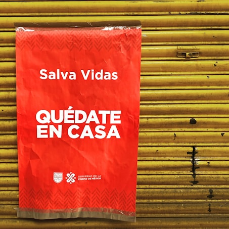 A bright red poster with white text that says 'Salva Vidas Qu&eacute;date En Casa' is attached to a yellow metal shutter. The message emphasizes staying at home to save lives. The bottom has logos representing the government of Mexico City.