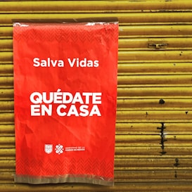 A bright red poster with white text that says 'Salva Vidas Qu&eacute;date En Casa' is attached to a yellow metal shutter. The message emphasizes staying at home to save lives. The bottom has logos representing the government of Mexico City.