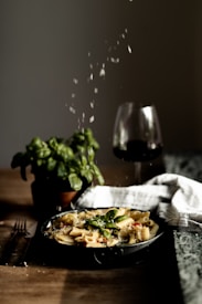 A plate of pasta garnished with basil leaves and grated cheese is placed on a wooden table. A potted plant and a glass of red wine are in the background, creating a cozy dining ambiance. The scene is warmly lit, highlighting the textures of the pasta and other elements.