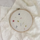 An embroidery hoop holds a piece of fabric with minimalist geometric designs in various colors, including red, yellow, blue, and gray. The hoop is placed on top of a larger piece of light-colored fabric.