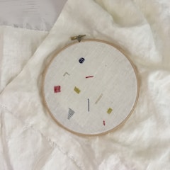 An embroidery hoop holds a piece of fabric with minimalist geometric designs in various colors, including red, yellow, blue, and gray. The hoop is placed on top of a larger piece of light-colored fabric.