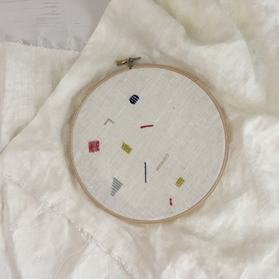 An embroidery hoop holds a piece of fabric with minimalist geometric designs in various colors, including red, yellow, blue, and gray. The hoop is placed on top of a larger piece of light-colored fabric.