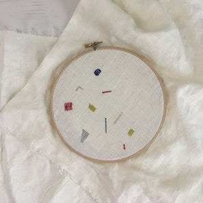 An embroidery hoop holds a piece of fabric with minimalist geometric designs in various colors, including red, yellow, blue, and gray. The hoop is placed on top of a larger piece of light-colored fabric.