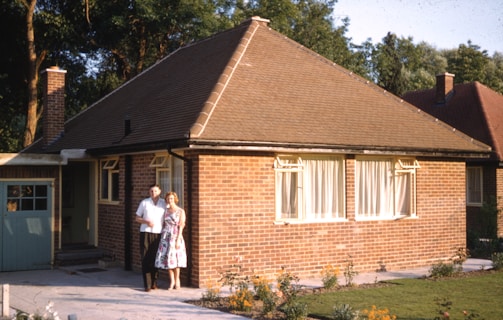 A quaint, single-story brick house with a triangular roof and a chimney stands surrounded by greenery. A couple stands in front of the house, with the man wearing a white shirt and the woman in a floral dress. The house has large windows with curtains, and a small garden with some flowers in bloom.