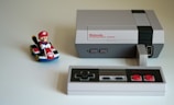 A classic Nintendo Entertainment System with a black and gray controller sits on a light surface. To the left of the console, there is a toy figure of a character in a red cap riding a small go-kart, adding a playful touch to the scene.