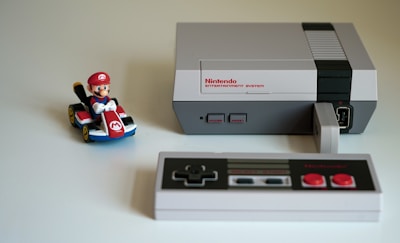 A classic Nintendo Entertainment System with a black and gray controller sits on a light surface. To the left of the console, there is a toy figure of a character in a red cap riding a small go-kart, adding a playful touch to the scene.