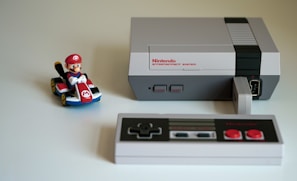A classic Nintendo Entertainment System with a black and gray controller sits on a light surface. To the left of the console, there is a toy figure of a character in a red cap riding a small go-kart, adding a playful touch to the scene.