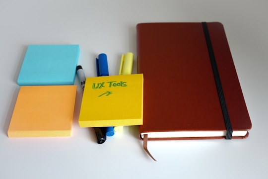 A collection of office supplies is arranged neatly, including colorful sticky notes in blue, orange, and yellow. The yellow note has 'UX Tools' written on it. There are various colorful markers and a thick brown notebook with an elastic band placed to the right.