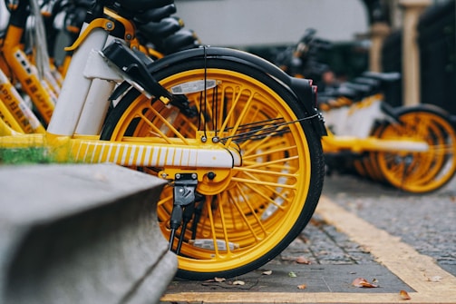 Bright yellow bicycle wheels are lined up in a row, partially in focus, with a close-up view of the spokes and tires. The pavement and curb are visible, with some blurred bicycles in the background.