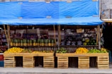 A vibrant fruit stand displays a variety of fresh fruits neatly arranged in wooden crates. The stand is covered with a blue tarp and features an array of pineapples, mangoes, oranges, and limes. In the background, packaged goods are visible, adding to the assortment of items available for sale.