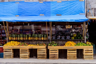 A vibrant fruit stand displays a variety of fresh fruits neatly arranged in wooden crates. The stand is covered with a blue tarp and features an array of pineapples, mangoes, oranges, and limes. In the background, packaged goods are visible, adding to the assortment of items available for sale.